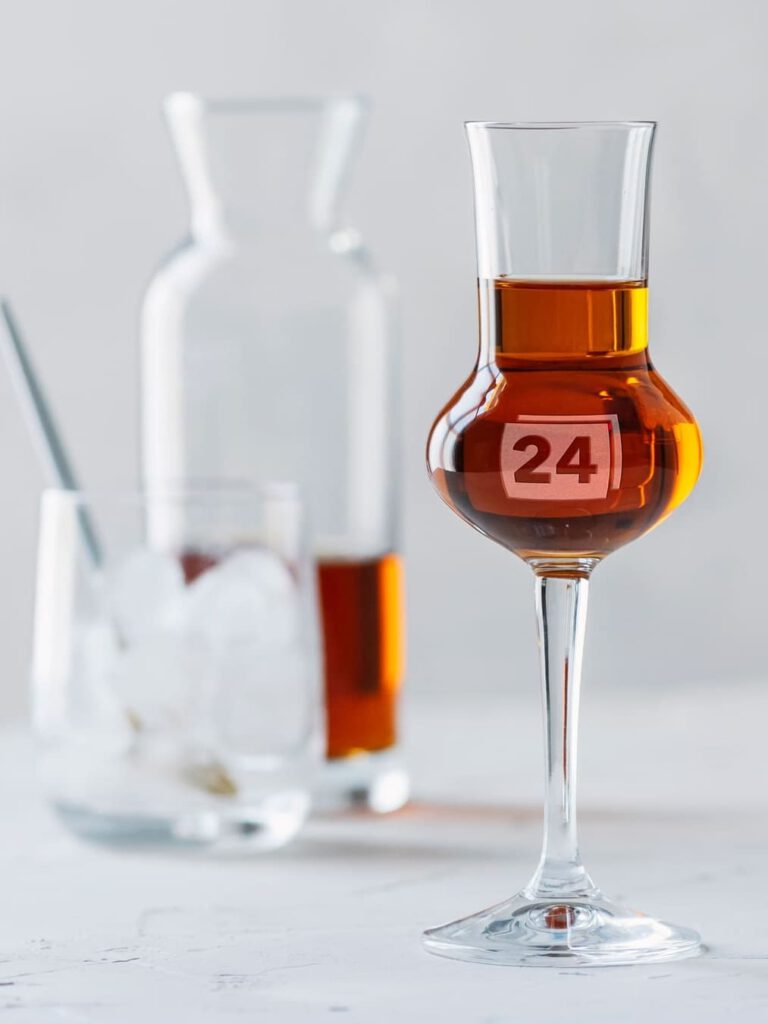 Grappaglas mit Gravur „24“ vor Karaffe und Glas mit Eiswürfeln