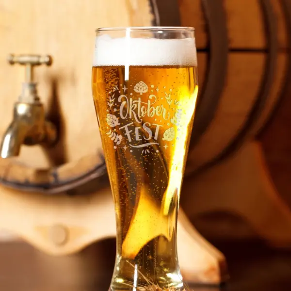 Weizenglas mit Gravur „Oktoberfest“, gefüllt mit Bier vor Holzfass