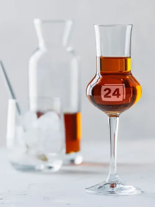 Grappaglas mit Gravur „24“ vor Karaffe und Glas mit Eiswürfeln